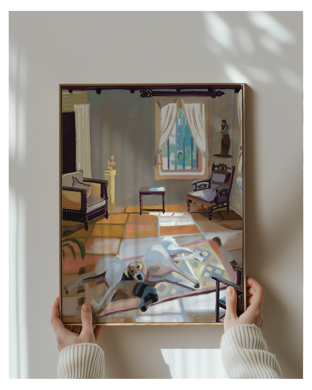 A framed art print depicting a stylized interior room with furniture and a dog, held by two hands wearing a cream ribbed sweater against a light gray wall with soft window shadows.
