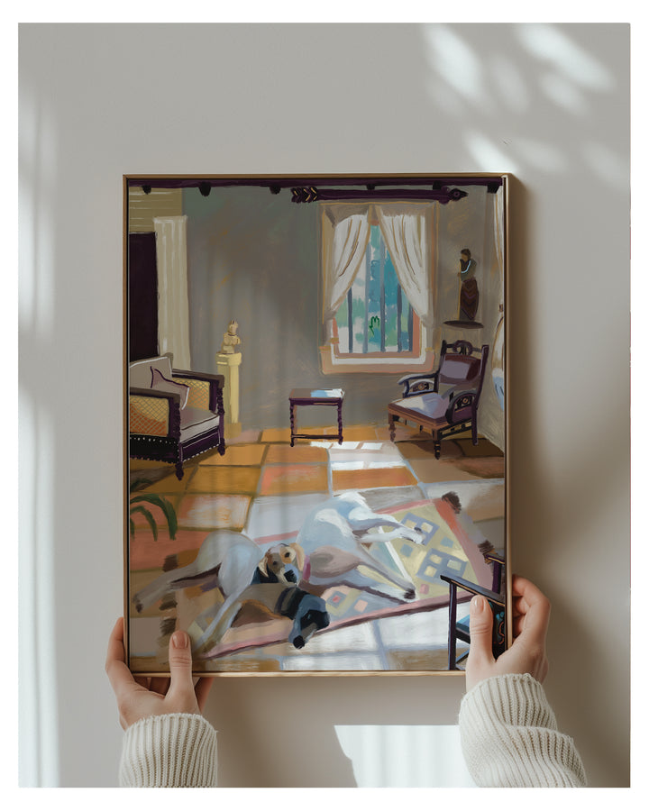 A framed art print depicting a stylized interior room with furniture and a dog, held by two hands wearing a cream ribbed sweater against a light gray wall with soft window shadows.