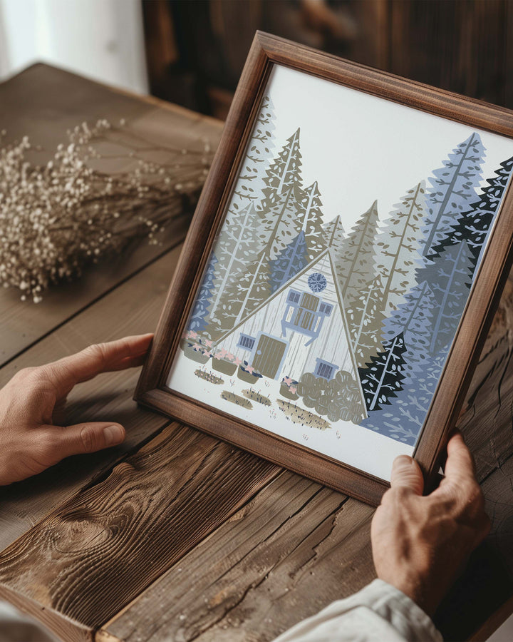 Person holding a Framed Artwork titled 'Alpine Retreat', featuring a charming A-frame cabin nestled among towering pines, created by little Berries Studio.