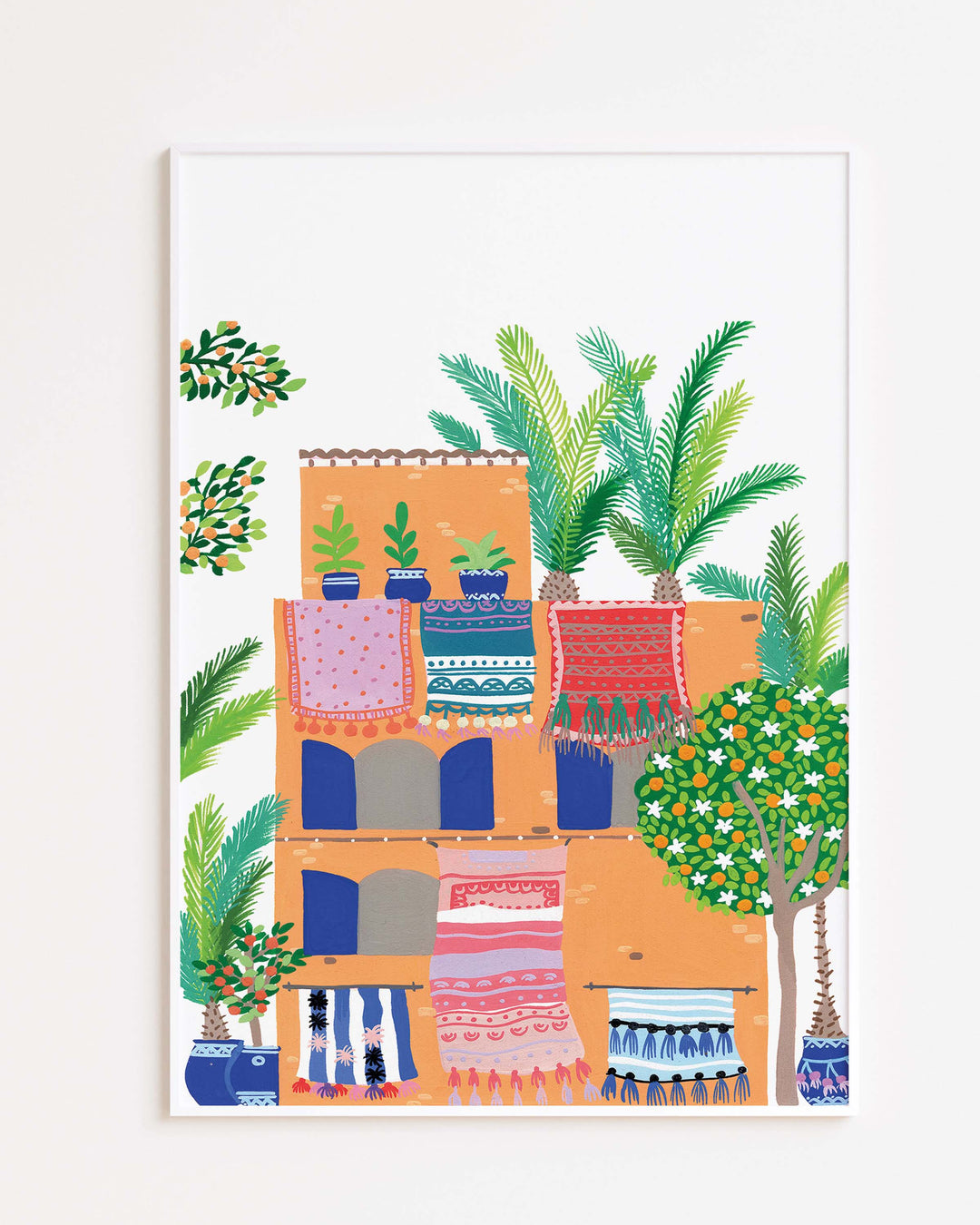 White Framed Artwork of a terracotta building with vibrant rugs hanging from balconies, surrounded by lush greenery and palm trees, by Little Berries Studio.