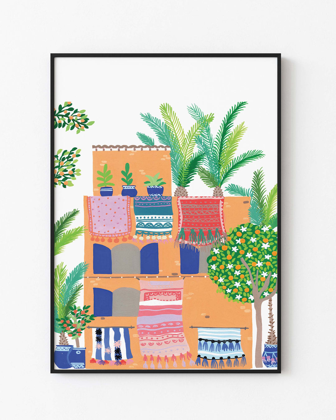 Black Framed Artwork of a terracotta building with vibrant rugs hanging from balconies, surrounded by lush greenery and palm trees, by Little Berries Studio.