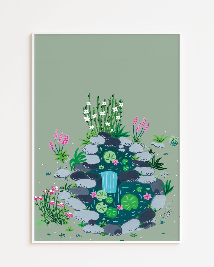 White Framed Art Print of a small, tranquil pond surrounded by smooth stones, blooming flowers, and lush greenery by Little Berries Studio.