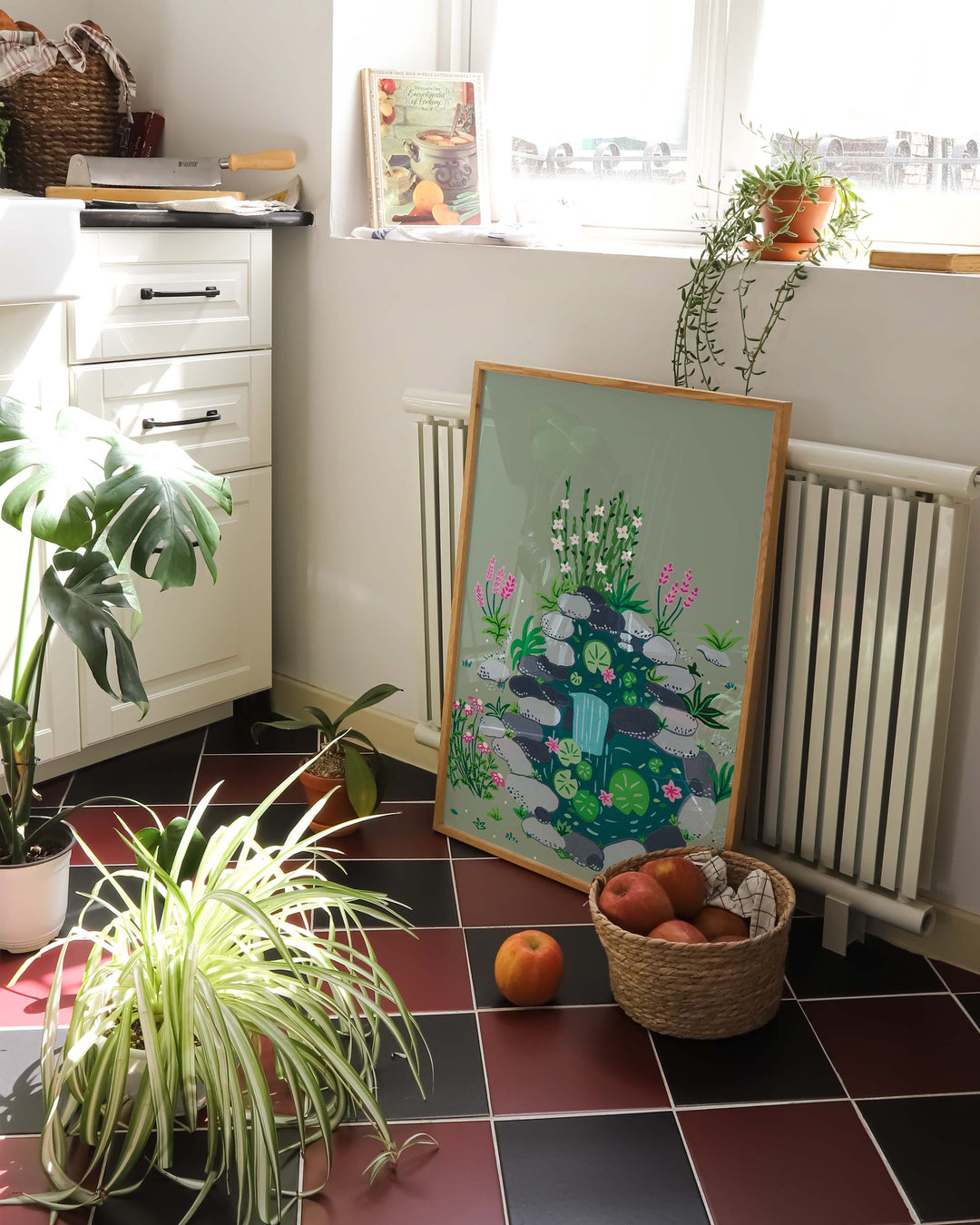 Framed Art Print of a tranquil pond scene with lily pads and blooming flowers, propped against a radiator in a bright, cozy kitchen filled with potted plants and a basket of apples on a tiled floor.