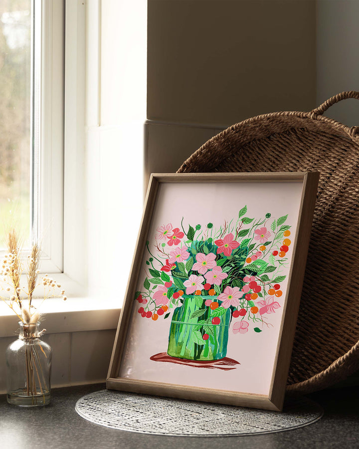 A framed Art Print by Little Berries Studio featuring a lush bouquet of pink flowers and green leaves in a green vase against a soft pink background, displayed on a table near a window with natural light.