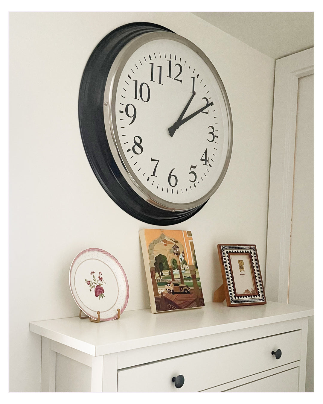 A large round wall clock with a black frame and white face, mounted on a white wall above a white dresser with decorative items.