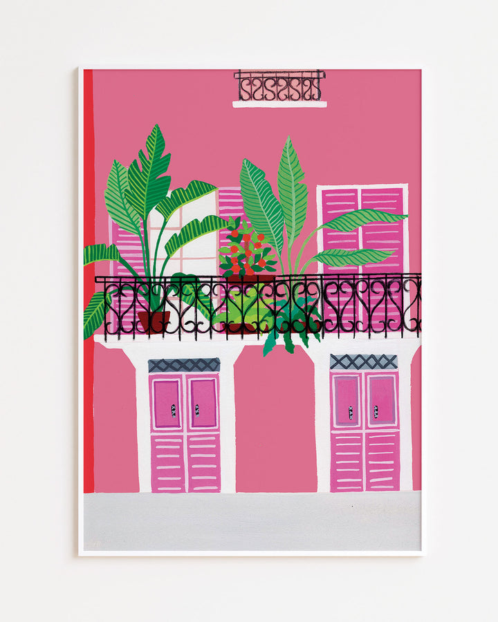 White Framed Artwork of a vibrant pink building with a black wrought-iron balcony adorned with potted tropical plants and bold pink shutters.