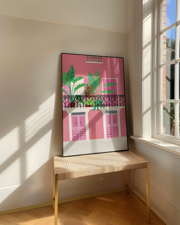 Framed Art Print titled 'Door Bloom Balcony' by Little Berries Studio, resting on a woven bench near a sunlit window in a bright, airy room.