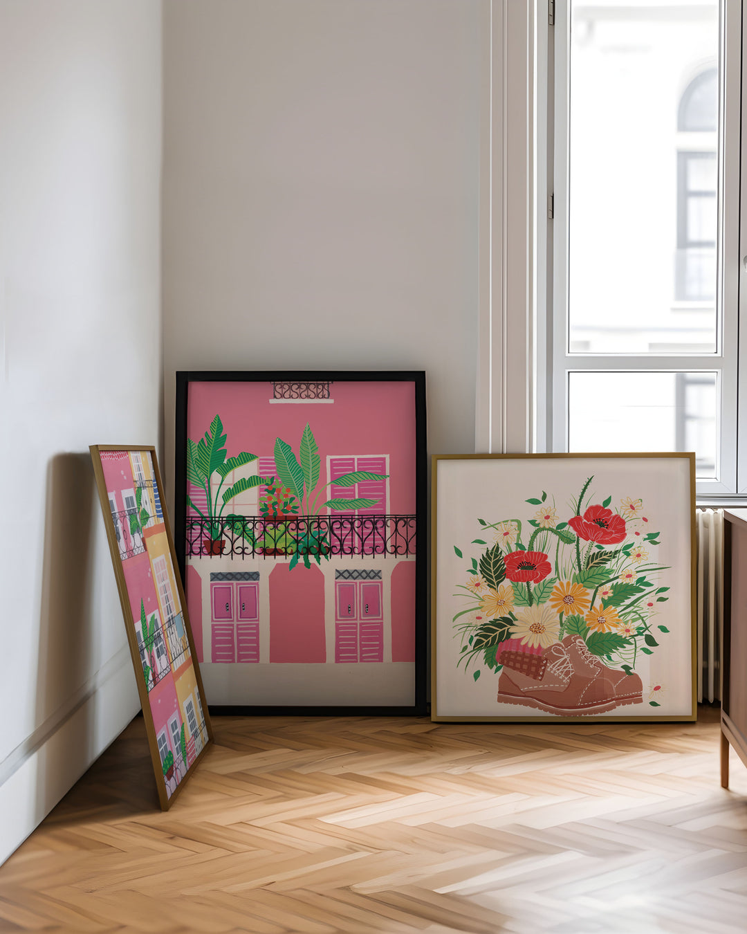 Art Prints created by Little Berries Studio leaning against a wall in a bright room, featuring a vibrant pink building with a tropical balcony scene and a floral composition with flowers growing out of a pair of shoes.
