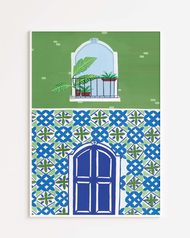 White Framed Artwork of a Mediterranean-style building with a vibrant blue door, patterned mosaic tiles, and a small balcony featuring tropical potted plants.