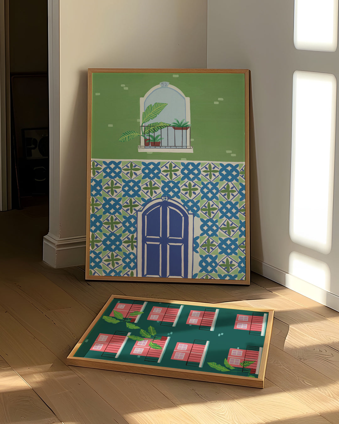 Art prints leaning against a wall in a bright room, showcasing a blue door framed by intricate mosaic tiles and a balcony with tropical plants and another Art Print showcasing pink doors and windows with tropical plants on balconies.
