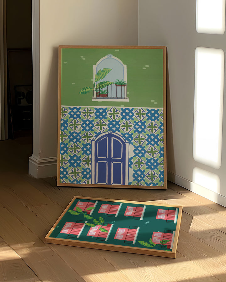 Art prints leaning against a wall in a bright room, showcasing a blue door framed by intricate mosaic tiles and a balcony with tropical plants and another Art Print showcasing pink doors and windows with tropical plants on balconies.