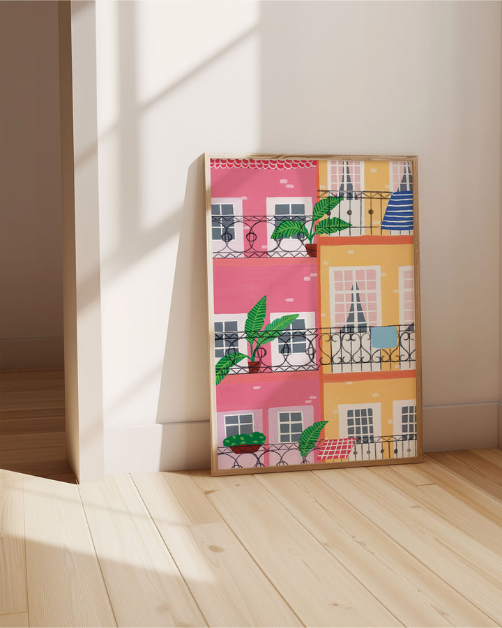 Framed Art Print titled "Door Urban Pastels" resting on the floor, featuring a vibrant illustration of colourful pastel buildings with balconies adorned by potted plants and decor.
