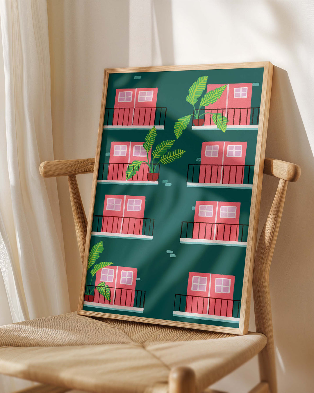 A framed Art Print leaning against a wooden chair, featuring a stylised illustration of a green building facade with pink doors and windows.