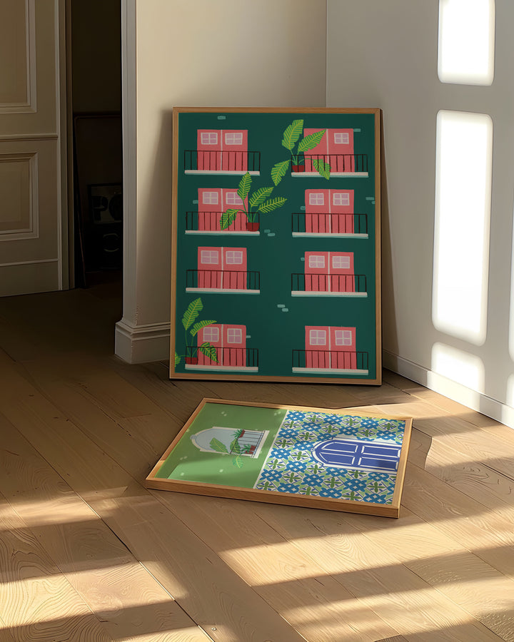 Art Print leaning against a wall in a bright room, showcasing a minimalistic design of pink doors and windows with tropical plants on balconies, another Art Print featuring a blue door framed by intricate mosaic tiles and a balcony with tropical plants rest on a floor.