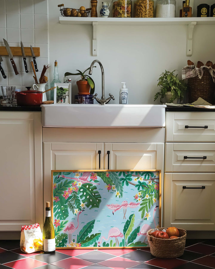Framed Art Print of a 'Flamingo Festivity' featuring pink flamingos wading in light blue water surrounded by lush green foliage, leaning against a kitchen island with a cozy, rustic decor including a basket of apples, wine bottle.