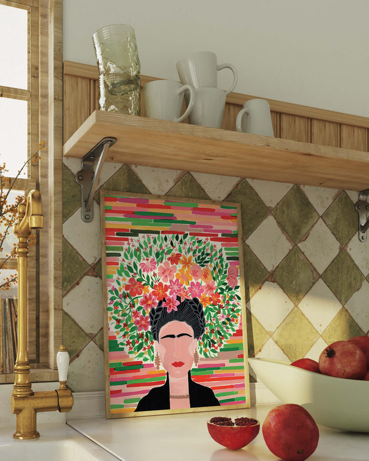 A framed botanical Art Print titled 'Floral Frida' featuring a portrait of Frida adorned with a lush floral crown resting on a kitchen shelf, created by Little Berries Studio.