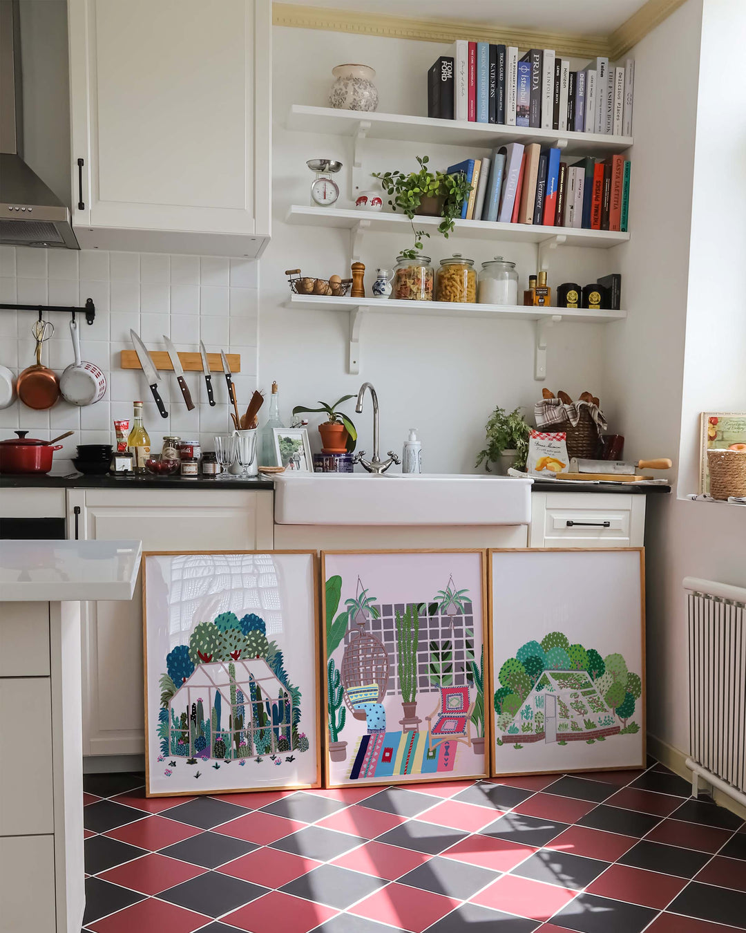 Photo of a cosy kitchen with white cabinetry, black and red checkered tile flooring, and open shelves filled with books, jars, and plants. Three colourful framed Art Prints are propped up against the cabinets, featuring greenhouse and plant-themed illustrations in vibrant colours.