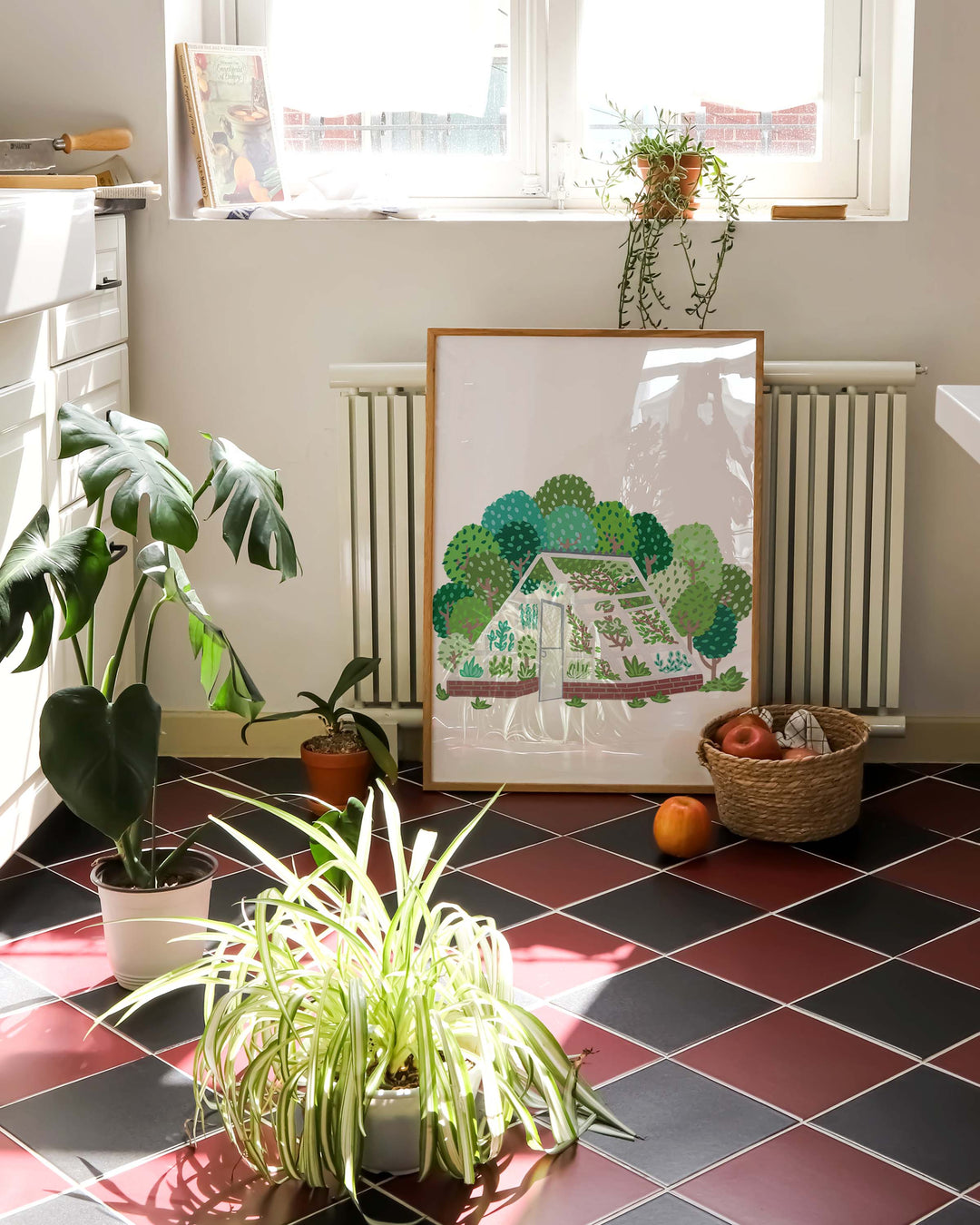 Art print titled 'Greenhouse Grove' by Little Berries Studio featuring a greenhouse illustration, displayed on the floor of a sunlit room with potted plants, a wicker basket, and a window in the background.