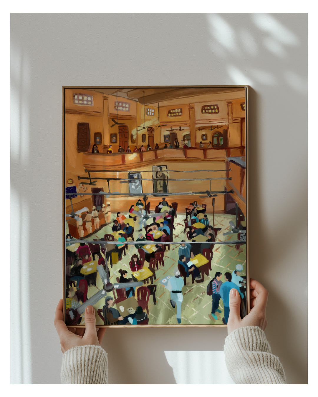 Framed artwork of a busy café scene held by two hands against a white wall.
