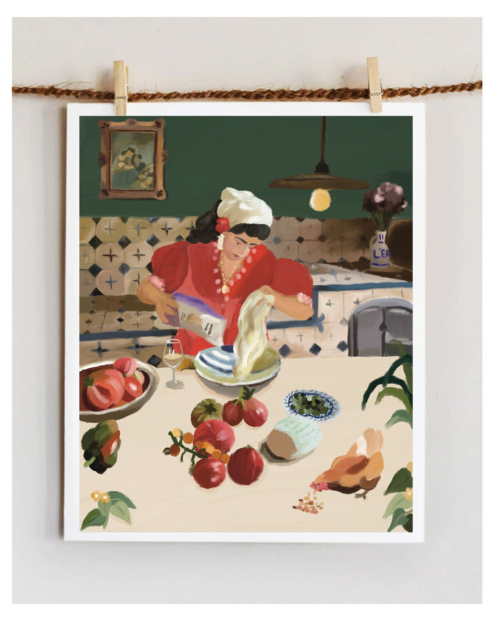 A framed art print depicting a woman in a red dress and white hat mixing dough at a table with fruit and a chicken, hung on a wall by two wooden clothespins.
