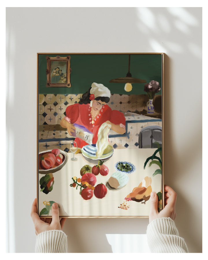 A framed art print depicting a woman in a red dress preparing food at a table with fruit and household items, held by two hands against a light gray wall.