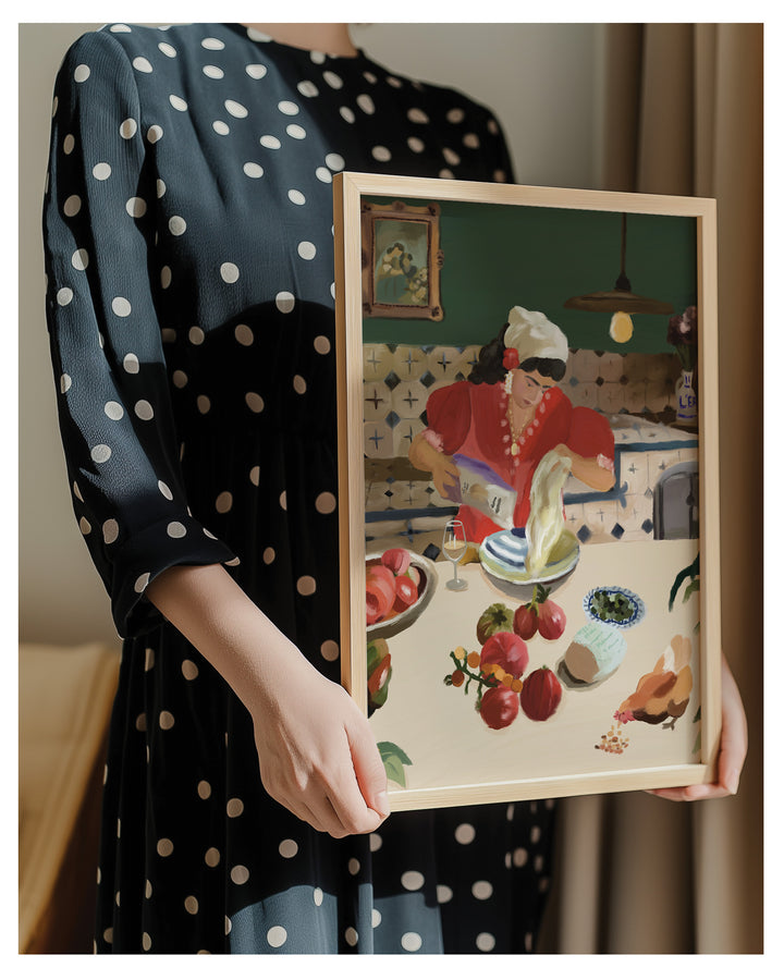 A person wearing a black dress with white polka dots holds a framed art print. The print features a colorful, stylized illustration of a woman in a kitchen preparing food at a table with fruit and dishes, set in a warm, cozy interior.