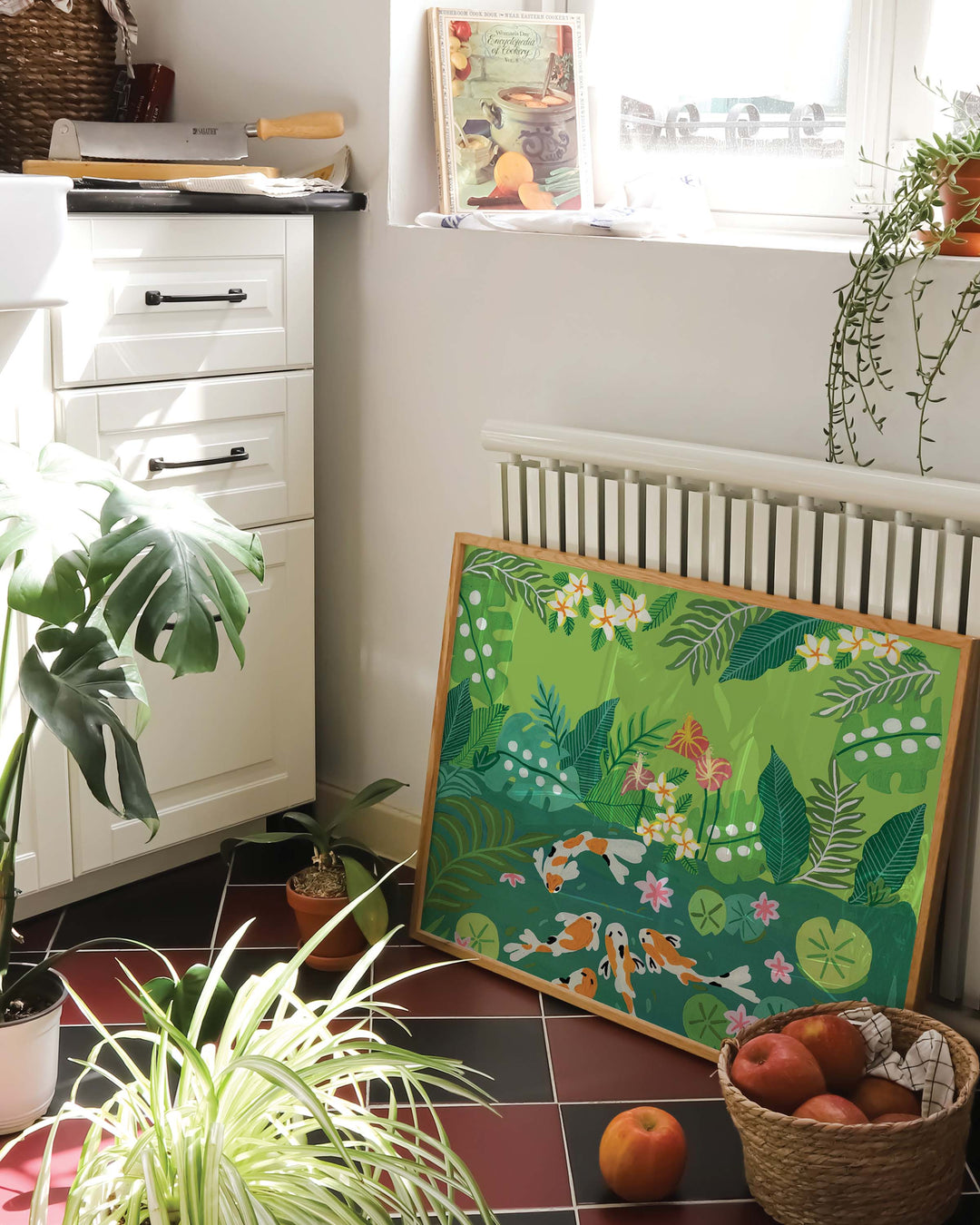 Framed Art Print of a koi fish swimming in a lush green garden pond surrounded by vibrant tropical foliage and blooming flowers, propped against a radiator in a bright, cozy kitchen filled with potted plants and a basket of apples on a tiled floor.