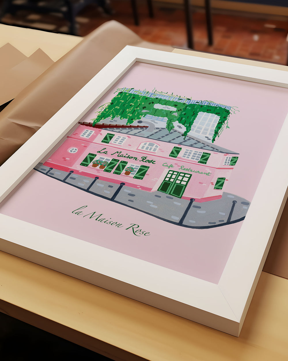 Framed Artwork of La’Maison Rose café in Montmartre, Paris, by Little Berries Studio, featuring a pink building with green shutters and lush green plants cascading from the roof.