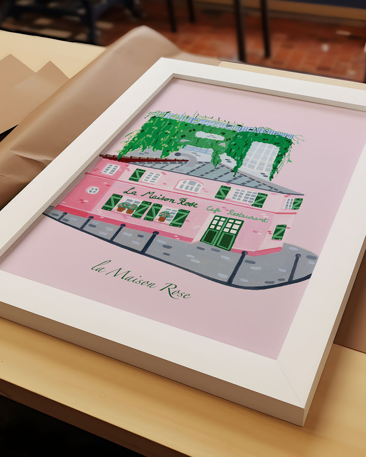 Framed Artwork of La’Maison Rose café in Montmartre, Paris, by Little Berries Studio, featuring a pink building with green shutters and lush green plants cascading from the roof.