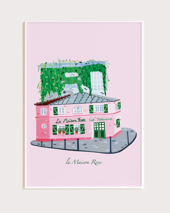 White Framed Artwork of La’Maison Rose café in Montmartre, Paris, by Little Berries Studio, featuring a pink building with green shutters and lush green plants cascading from the roof.
