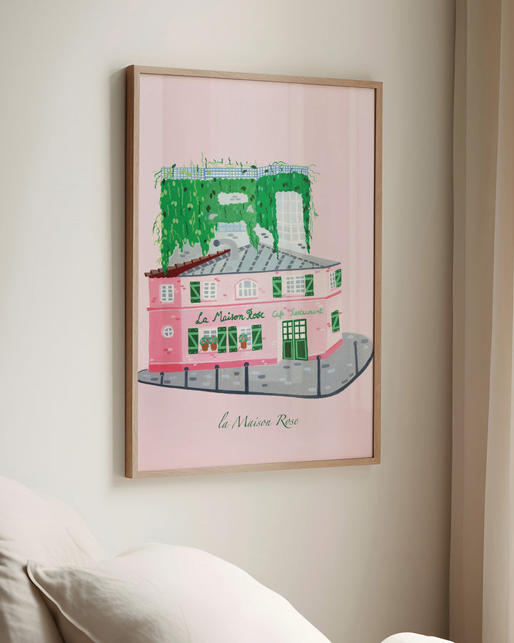 Framed Wall Art of La’Maison Rose café in Montmartre, Paris, by Little Berries Studio, featuring a pink building with green shutters and lush green plants cascading from the roof.