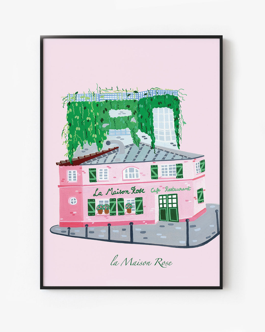 Black Framed Artwork of La’Maison Rose café in Montmartre, Paris, by Little Berries Studio, featuring a pink building with green shutters and lush green plants cascading from the roof.