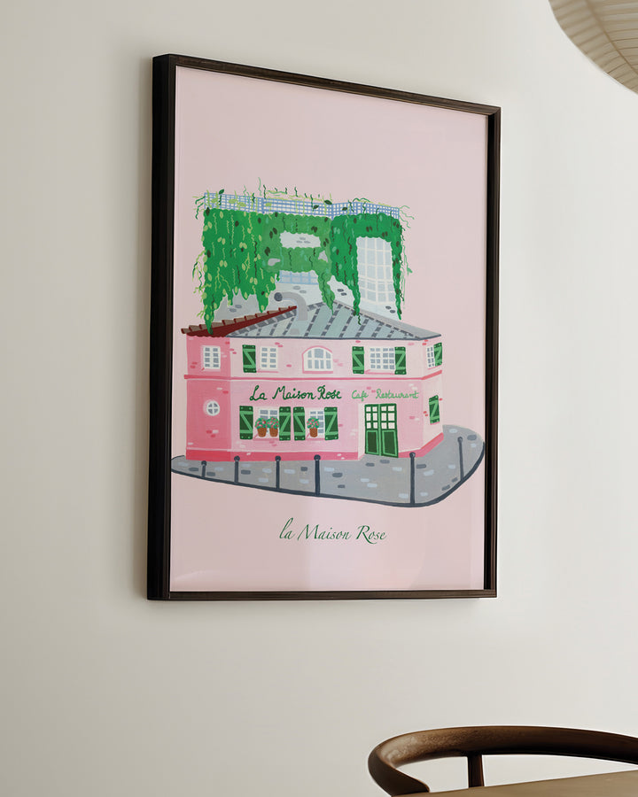 Ready-to-Hang Framed Wall Art of La’Maison Rose café in Montmartre, Paris, by Little Berries Studio, featuring a pink building with green shutters and lush green plants cascading from the roof.