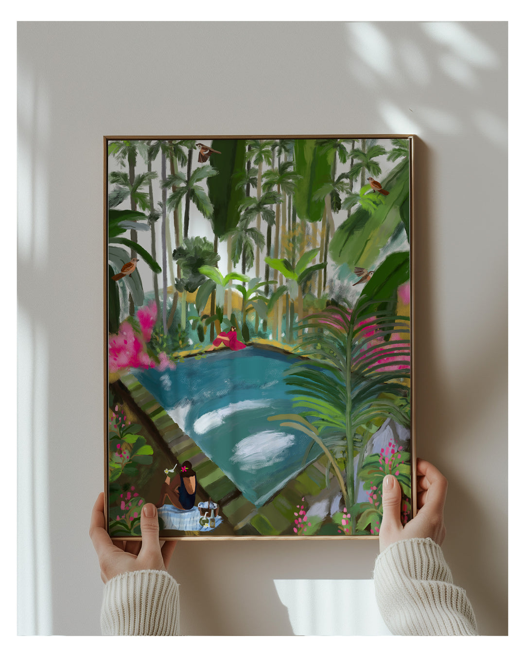 Framed artwork of a tropical scene with a pond and people, held by two hands.