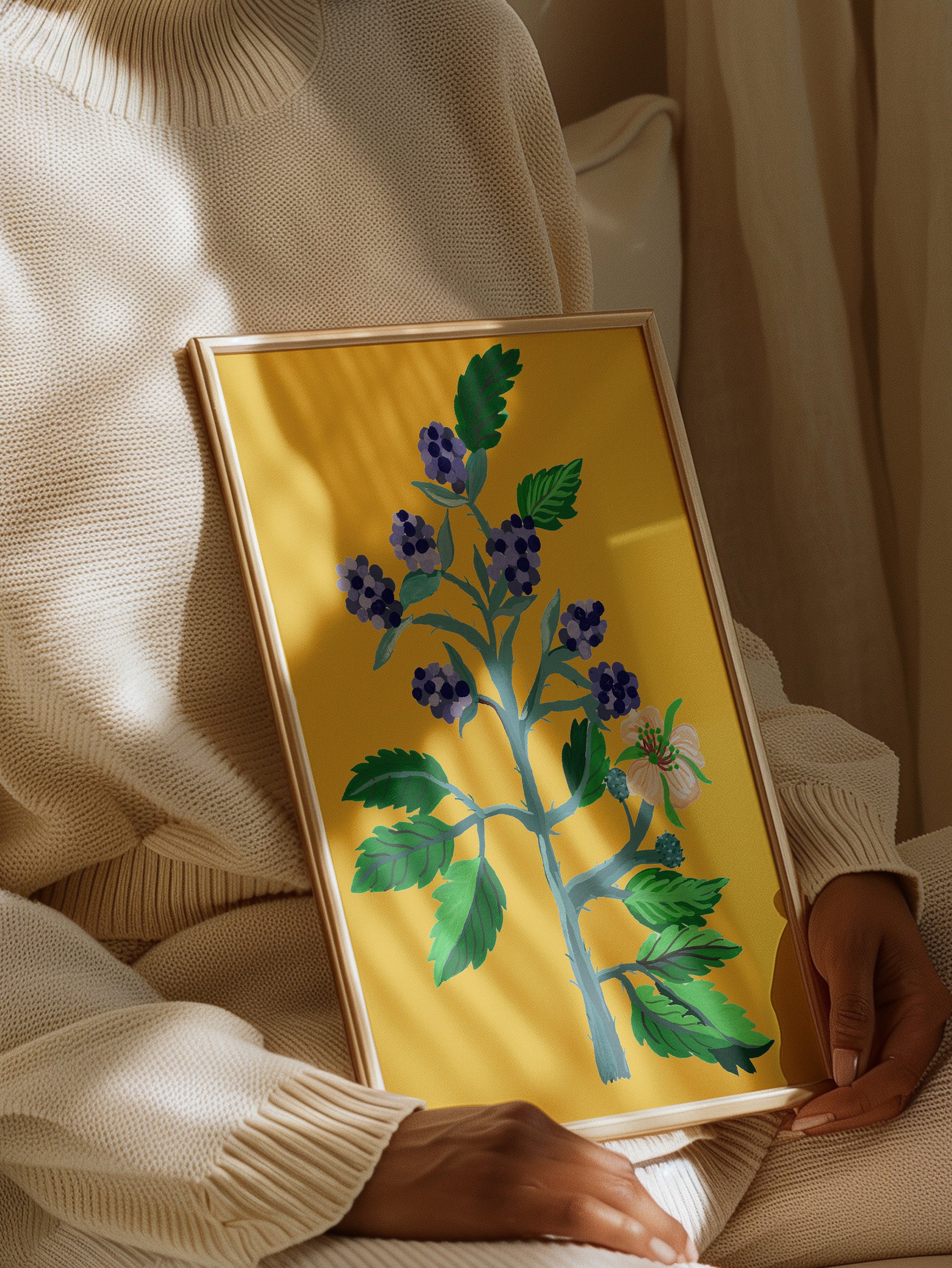 A framed botanical Art Print featuring vibrant blue berries on a yellow background. The image shows the Print being held by a person in a cosy, cream-coloured outfit.