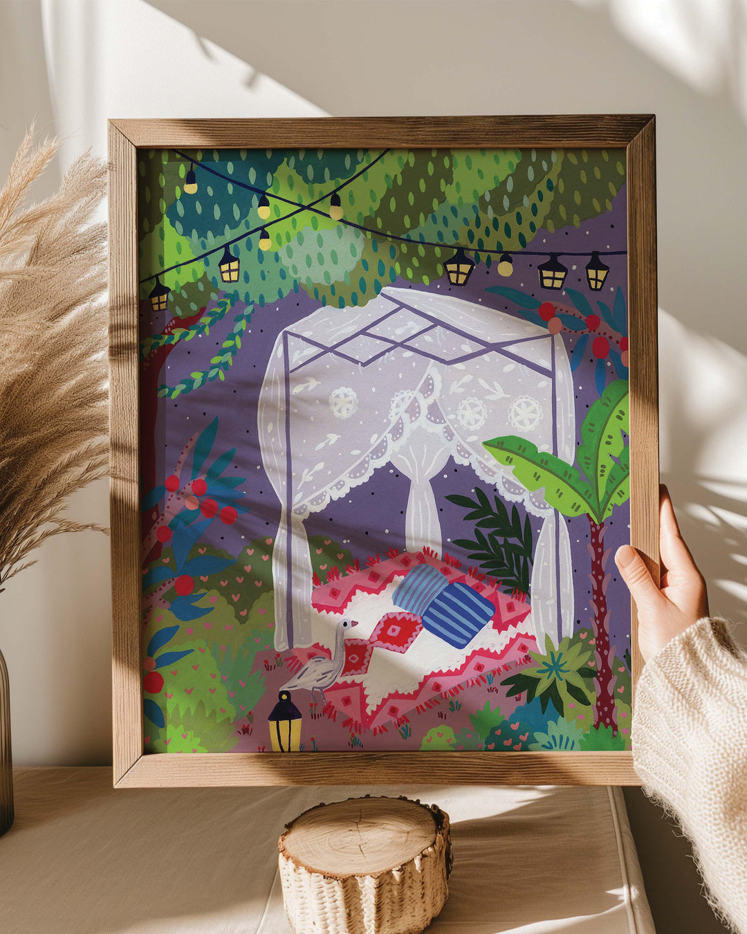 A framed Art Print being held by a person, featuring a whimsical outdoor scene with a lace-draped canopy bed under a canopy of trees. The bed is set on a vibrant patterned blanket with pillows, surrounded by lush greenery, palm trees, and glowing string lights overhead.