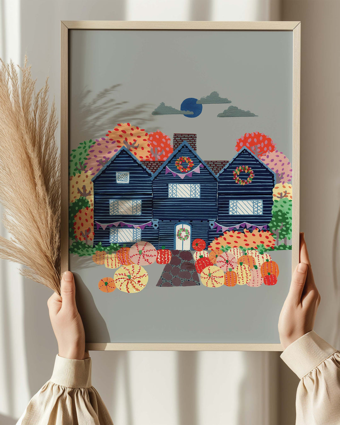 Person holding an Artwork of a cosy, orange Victorian house surrounded by trees, capturing the essence of autumn with festive details and warm seasonal colours.
