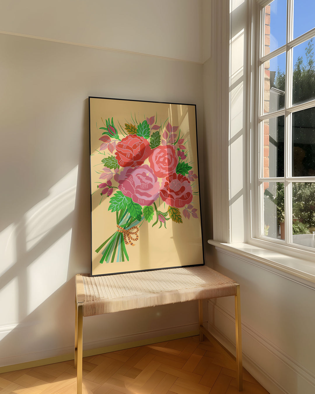 Framed Art Print titled 'Rose Reverie' by Little Berries Studio, resting on a woven bench near a sunlit window in a bright, airy room.