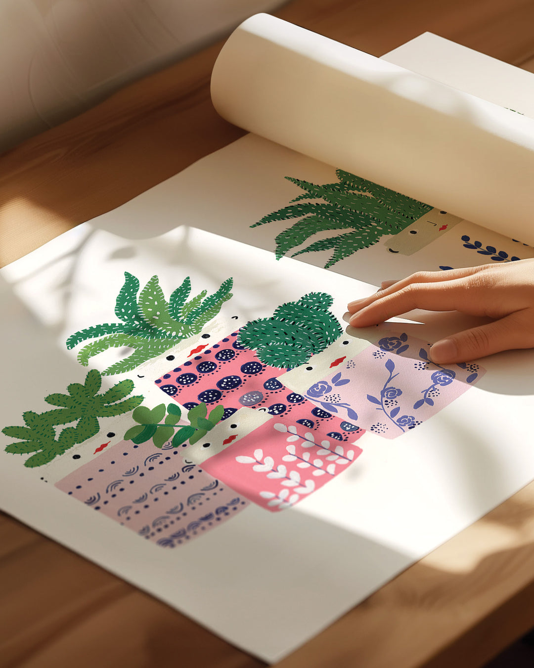 A close-up of a person’s hand touching an Art Print depicting several potted succulents. The print is unrolled on a wooden surface, with soft sunlight casting shadows across the scene, creating a warm and inviting atmosphere.
