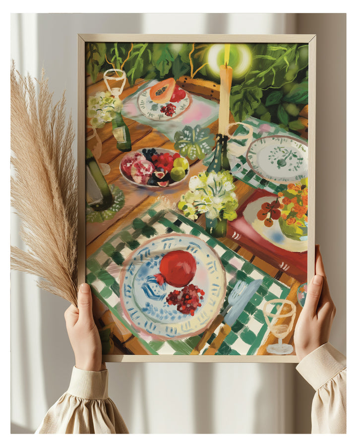 A framed Art print depicting a colorful still life table scene with plates, fruit, flowers, and candles, held by two hands against a soft beige background with dried grass on the left.