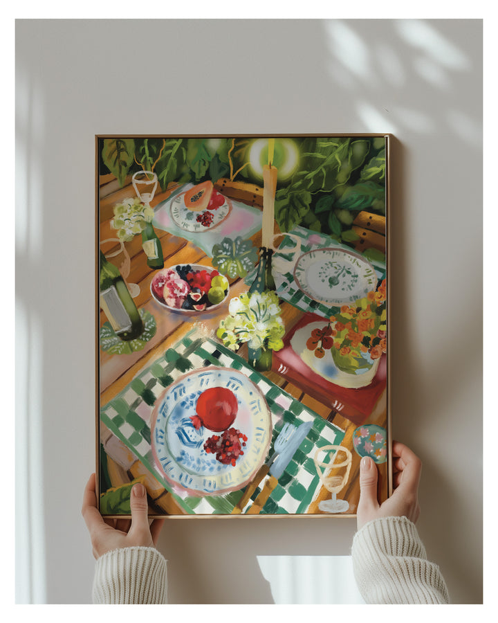 A framed still life painting held up by two hands against a light gray wall with soft window shadows. The artwork depicts a colorful dining table scene with plates, flowers, fruit, and green foliage.