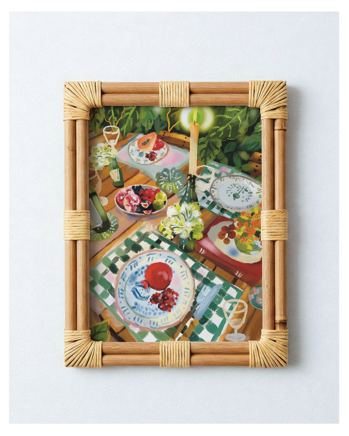 A framed art print featuring a colorful still life painting of a table setting with plates, flowers, and greenery, enclosed in a natural rattan frame against a light gray background.
