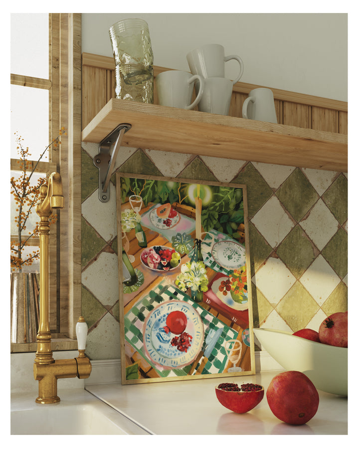 A framed art print featuring a colorful vintage-style table setting with fruit and plates, leaning against a tiled kitchen wall next to a brass faucet and pomegranates.
