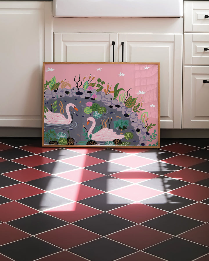 Framed Art Print of a 'Swanlake Whispers' featuring a tranquil scene of two pink swans gliding gracefully across a serene pond, surrounded by lush greenery, lily pads, and soft pink flowers leaning against a kitchen island with a cozy and rustic decor.