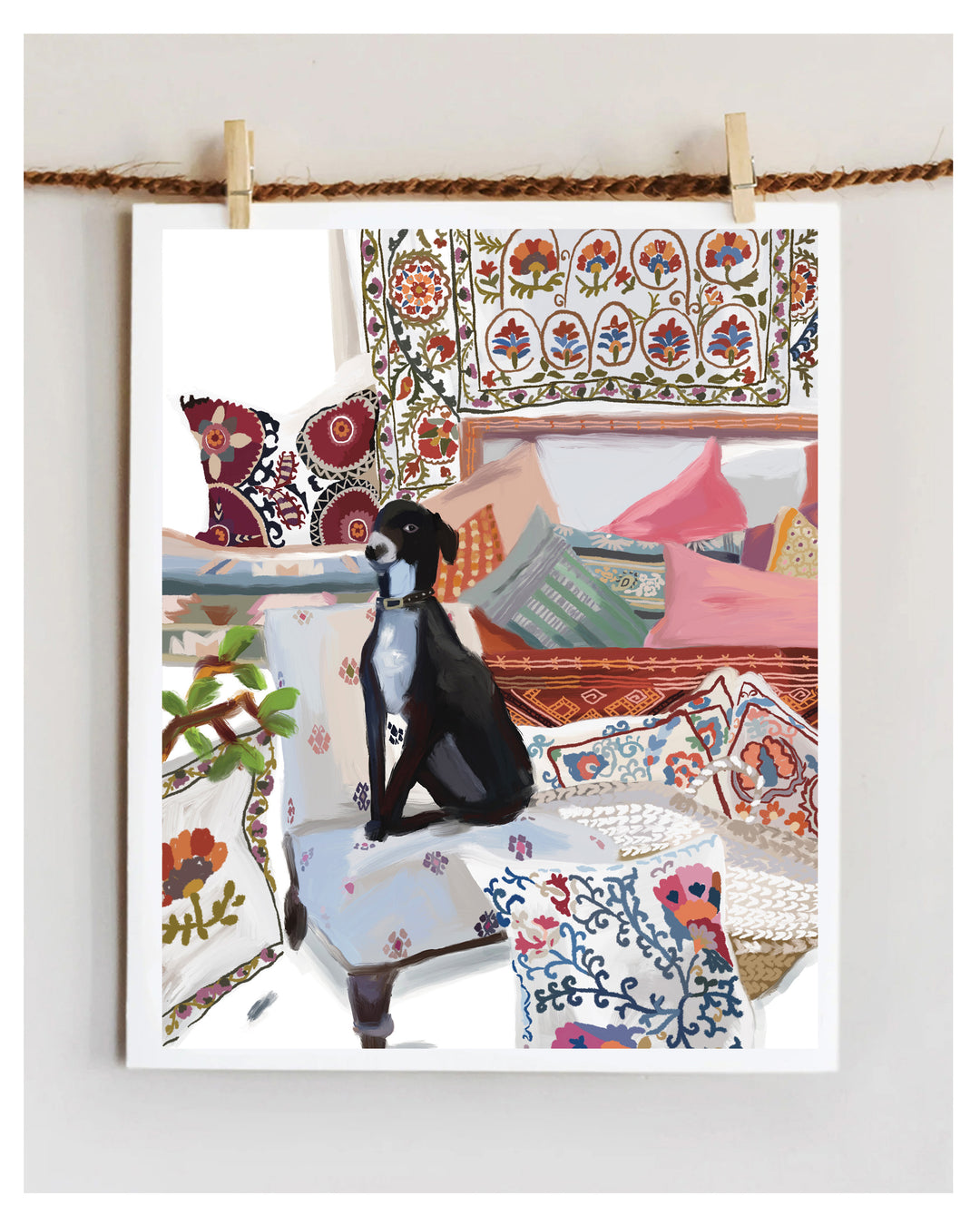 A framed art print depicting a black and white dog sitting in a cozy, patterned room with colorful cushions and textiles, hanging from a rope with wooden clips against a light wall.