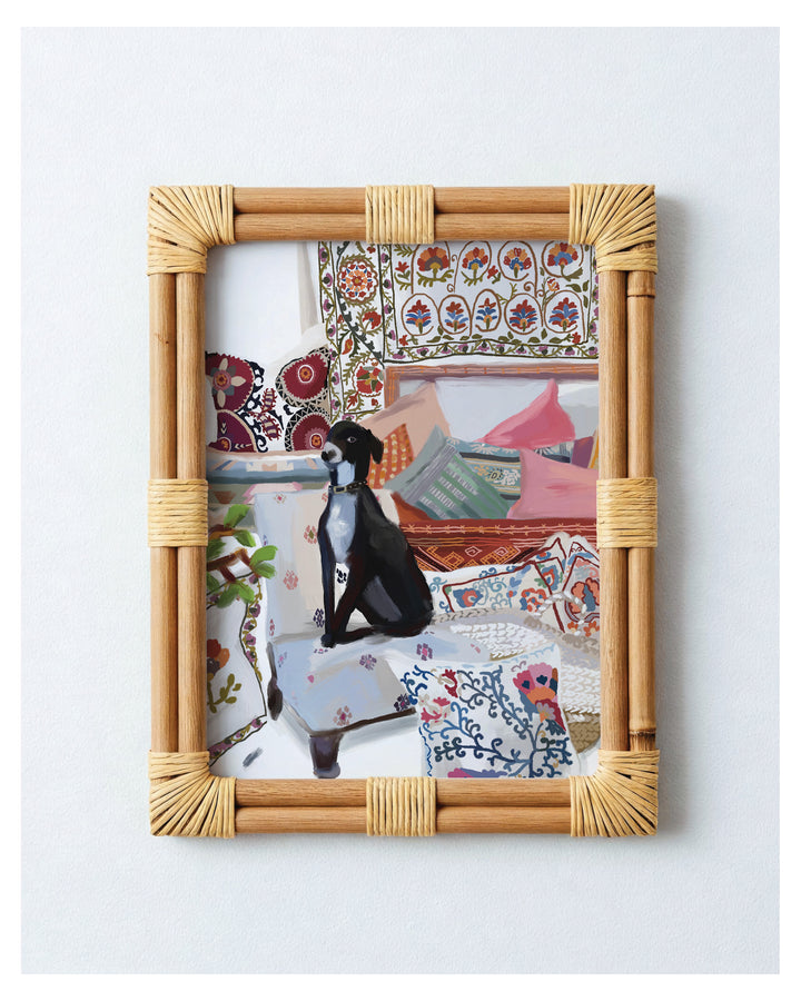 A framed art print featuring a black and white dog sitting on a patterned sofa in a colorful, bohemian-style room. The print is housed in a light bamboo frame with natural fiber wrapped corners, displayed against a clean white wall.