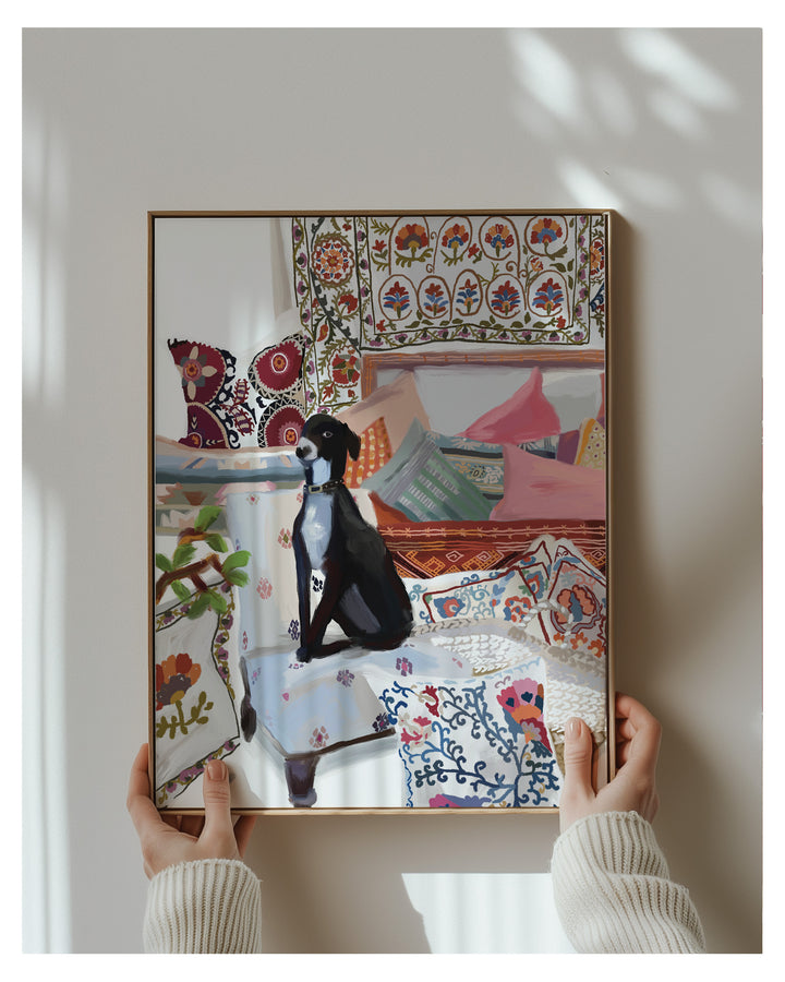 A framed art print depicting a black and white dog sitting amidst a colorful, patterned room filled with pillows, held by two hands wearing a cream ribbed sweater against a neutral wall with soft shadows.