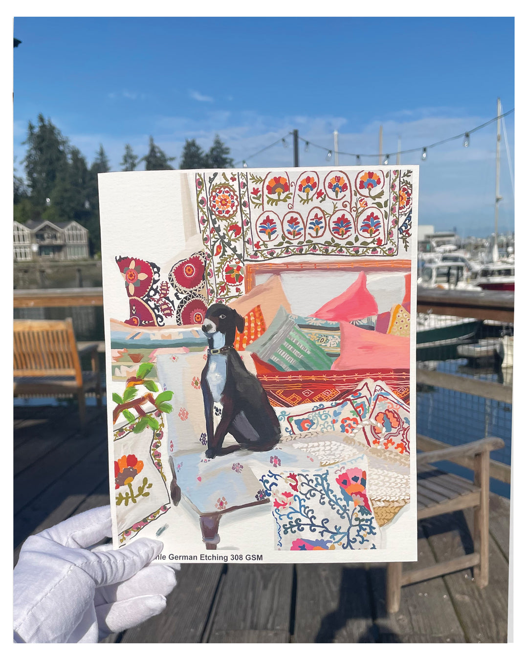 A hand wearing a white glove holds a printed art print featuring a colorful illustration of a black dog in a bohemian-style room with patterned textiles. The background shows a real outdoor harbor scene with boats, water, and wooden dock furniture.