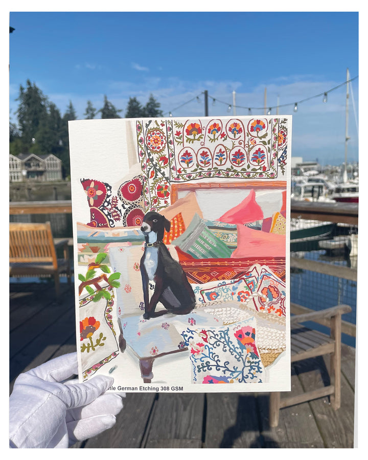 A hand wearing a white glove holds a printed art print featuring a colorful illustration of a black dog in a bohemian-style room with patterned textiles. The background shows a real outdoor harbor scene with boats, water, and wooden dock furniture.