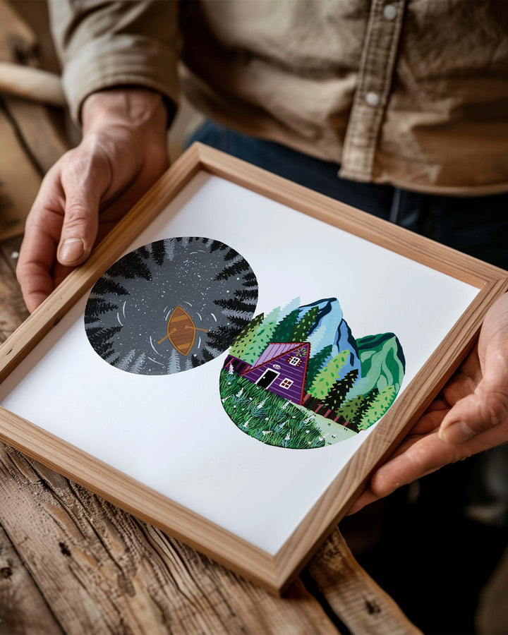 Person holding a Framed Artwork of two circular nature illustrations: one showing a boat in a dark lake surrounded by trees at night, the other with a cosy cabin in the mountains with lush greenery.